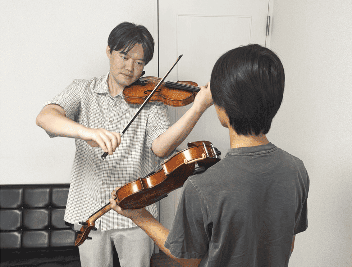 レッスン風景 講師が男子の前でヴァイオリンを弾いて見せている場面
